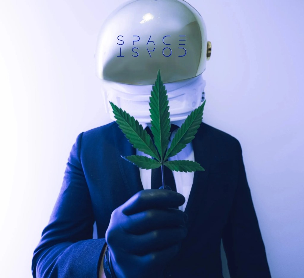 A person in a suit and gloves wearing a gold astronaut helmet, holding a cannabis leaf in front of a light blue background.