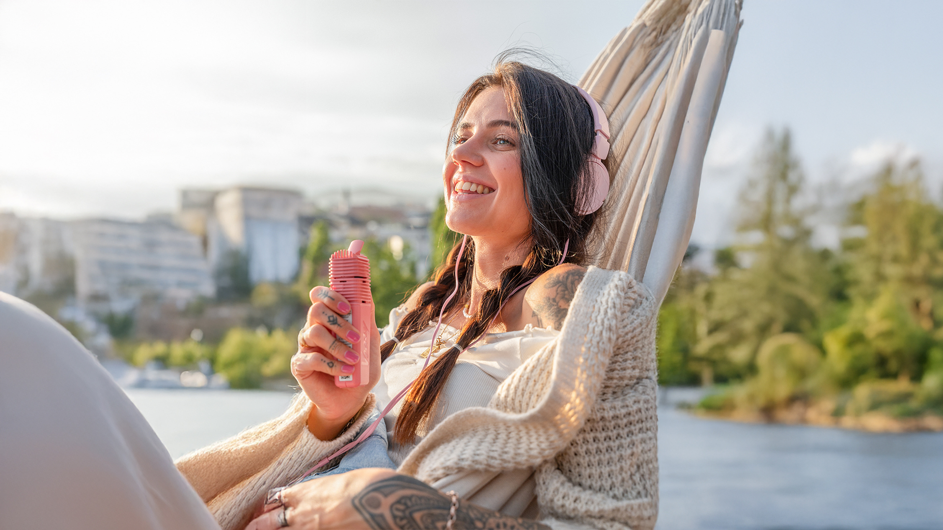 Smiling woman wearing pink headphones, relaxing in a hammock by water and holding a pink Storz & Bickel Venty vaporizer